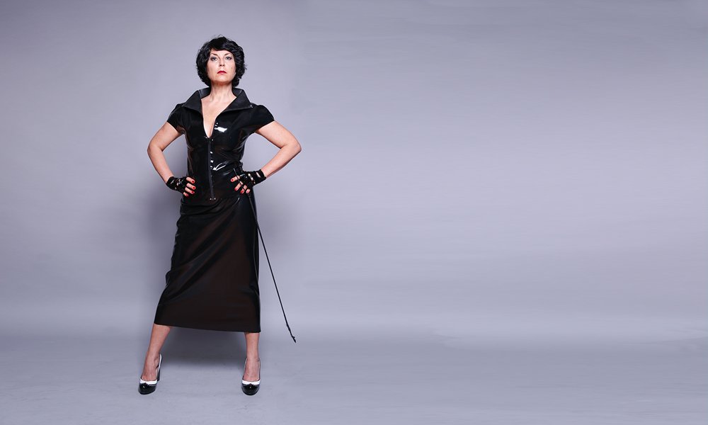 Domina Linda Dorn aus Frankfurt in schwarzer Latex Bluse und langem schwarzem Rock streng mit Gerte stehend